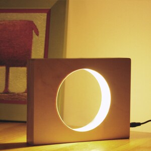 May include: A wooden lamp with a circular cutout that emits a warm, white light. The lamp is a light brown colour and has a simple, modern design.