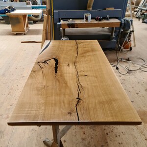 Könnte beinhalten: Eine große, hellbraune Tischplatte aus Holz mit natürlicher Holzmaserung und einem dunklen Riss, der in der Mitte verläuft. Die Tischplatte wird von einem einfachen Metallgestell getragen.