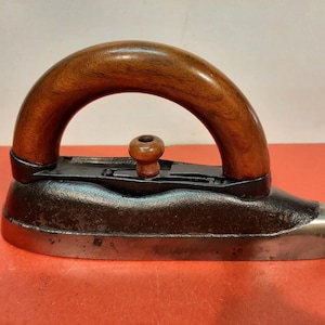 Pode incluir: Um ferro de engomar vintage com uma base de ferro fundido escuro e texturizado e uma base de metal polido. O cabo é feito de madeira castanha lisa e arqueada, complementado por um pequeno botão central de madeira para ajustar o mecanismo.