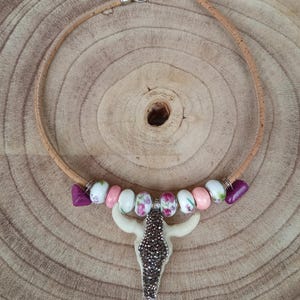Peut inclure: Un collier avec un cordon en liège beige et un fermoir argenté. Le pendentif est une tête de taureau blanche avec un centre scintillant marron. Le collier a des perles roses, blanches et violettes.