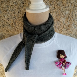 Peut inclure: Une écharpe tricotée gris foncé est drapée autour du cou d'un mannequin, avec les extrémités pendantes. Une broche décorative représentant une poupée en robe rose est épinglée sur le t-shirt blanc. L'arrière-plan est une surface texturée brun clair.