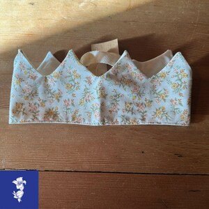 May include: A fabric crown with a floral pattern in pastel colors. The crown features a light blue background with pink and yellow flowers. A cream-colored ribbon is attached for securing the crown. The crown is approximately 10 inches wide.