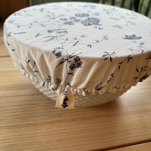 May include: A white bowl with a floral patterned fabric cover. The cover features a blue floral design and is gathered around the bowl's rim. A small tag with a blue floral design is attached to the cover.