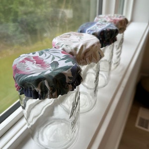 Peut inclure: Quatre bocaux en verre avec des couvercles en tissu. Les couvercles sont à motifs floraux avec des couleurs rose, vert et blanc. Les bocaux sont alignés sur un rebord de fenêtre blanc.