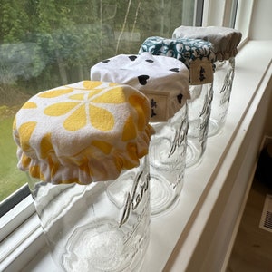 Peut inclure: Quatre bocaux en verre avec des couvercles en tissu. Les couvercles sont de différents motifs, notamment des fleurs jaunes, des cœurs noirs, des feuilles vertes et des pois gris. Les bocaux sont en verre transparent et portent une étiquette blanche sur laquelle est inscrit "Mason Jars".