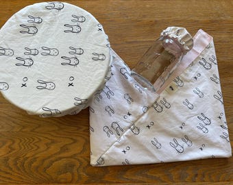 May include: Two white fabric food covers with a black bunny rabbit pattern. One cover is round and the other is a square with pink trim. The square cover is folded and a clear glass jar is partially visible.