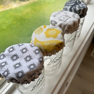 Peut inclure: Quatre bocaux en verre avec des couvercles en tissu. Les couvercles ont des motifs différents : des soleils noirs et blancs, des fleurs jaunes, un chat noir et blanc et un motif gris et blanc. Les bocaux sont étiquetés "Kerr" et sont posés sur une surface blanche.