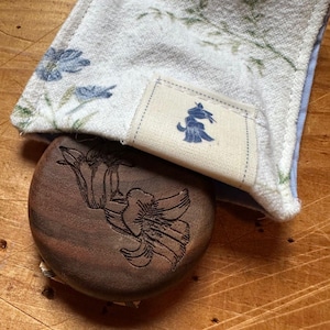 Peut inclure: Une boîte à bijoux ronde en bois avec un motif floral gravé sur le couvercle. La boîte est recouverte d'un tissu blanc à motif floral bleu et d'une petite étiquette en tissu bleu avec une illustration stylisée d'un chien.