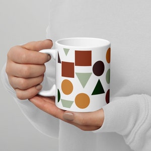 Pode incluir: Caneca de cerâmica branca com um padrão geométrico em tons de marrom, verde, laranja e vermelho. O padrão inclui círculos, quadrados e triângulos.