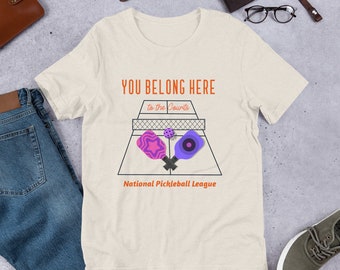 Może przedstawiać: Beżowa koszulka z napisem "YOU BELONG HERE to the courts" i grafiką przedstawiającą dwie rakiety do pickleballa i piłkę na korcie. Pod grafiką znajduje się napis "National Pickleball League".