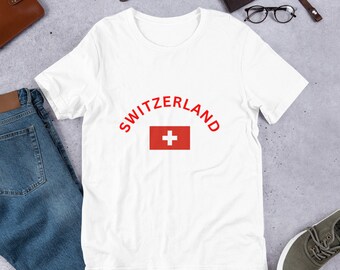 Può includere: Maglietta bianca con una grafica rossa della bandiera svizzera e la parola "SWITZERLAND" in rosso.