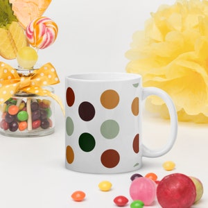 Puede incluir: Taza de cerámica blanca con un patrón de lunares en tonos marrones, verdes y beige. La taza tiene un asa redondeada.