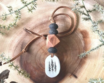 May include: A brown leather cord necklace with a white ceramic pendant featuring a black silhouette of three pine trees. The necklace also includes a brown wooden bead and black lava rock beads.