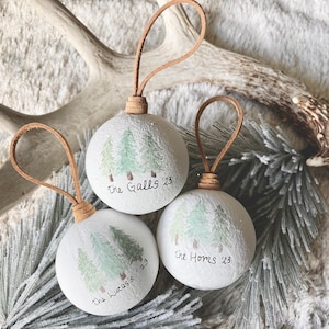 May include: Three white round ornaments with a brown leather cord. Each ornament has a watercolor painting of three green pine trees and the text "The Galls '23", "The Homes '23", and "The Lucas' '23".