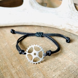 May include: A black leather bracelet with a silver gear charm. The bracelet is adjustable and has a simple design.