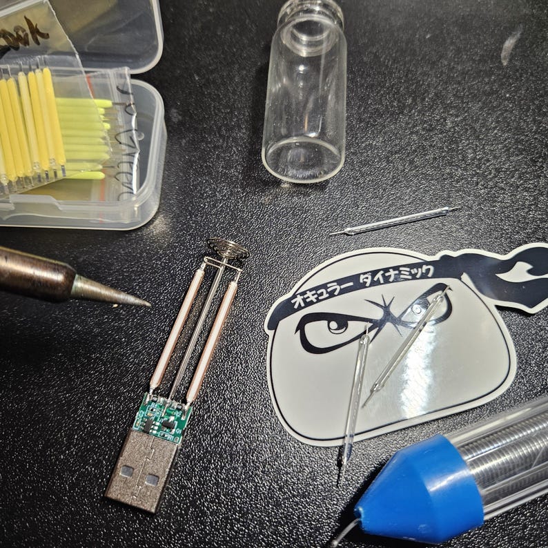 May include: An assortment of electronic components and tools, including a soldering iron, a clear glass vial, and a USB connector with attached wires. A sticker with Japanese text and a cartoon eye design is also present, along with a container of yellow and clear components.
