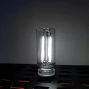 May include: A clear glass candle holder with a thick, round base and a cylindrical body. Two vertical, bright white lights are visible inside the holder, creating a glowing effect. The holder sits on a dark surface.
