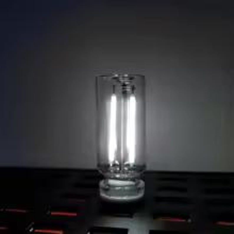 May include: A clear glass candle holder with a thick, round base and a cylindrical body. Two vertical, bright white lights are visible inside the holder, creating a glowing effect. The holder sits on a dark surface.