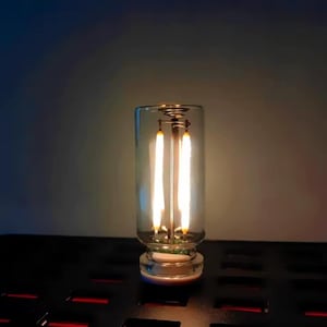 May include: A clear glass light bulb with two vertical filaments. The bulb has a cylindrical shape and a decorative base. The filaments are lit, emitting a warm, yellow glow. The bulb is designed for decorative lighting.