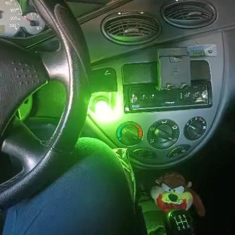 May include: Interior view of a car dashboard with a steering wheel, radio, and air conditioning controls. A Taz plush toy sits near the gear shift. The dashboard is illuminated with a green glow.