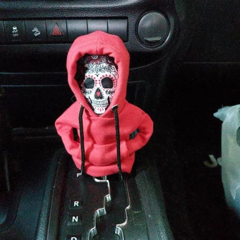 May include: A Day of the Dead sugar skull figure wearing a red hoodie with black drawstrings. The skull has intricate floral and geometric designs in white, red, and black. The figure is positioned in a car.