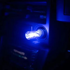 May include: A glowing blue LED light bulb is inserted into a car's dashboard. The bulb emits a bright, cool blue light, contrasting with the dark interior. The image is a close-up, highlighting the bulb's illumination and the surrounding details.