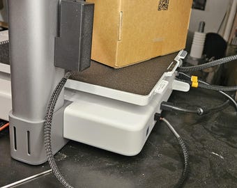 Può includere: Primo piano di una stampante 3D con un alimentatore nero attaccato al lato. La stampante è su una piattaforma bianca con un cavo nero che va dall'alimentatore alla stampante. La stampante è su una superficie nera. La stampante ha una scatola di cartone marrone sopra. La scatola ha un codice QR e il testo "Per qualsiasi problema con il filamento, scansiona il codice QR per accedere allo strumento ufficiale del filamento."