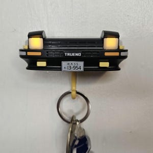 May include: Black car-shaped key holder with the word "TRUENO" and a Japanese license plate. The key holder has a yellow hook and is mounted on a white wall. The headlights are illuminated.