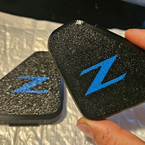 May include: Two black triangular car emblems with a blue "Z" logo. The emblems have a textured surface.