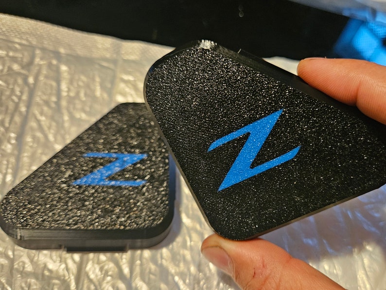 May include: Two black triangular car emblems with a blue "Z" logo. The emblems have a textured surface.
