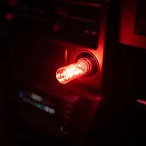 May include: A close-up of a car's interior, illuminated by a bright red light. The light source is a cylindrical object inserted into a circular socket, casting a warm glow against the dark surroundings. The dashboard and console are visible.
