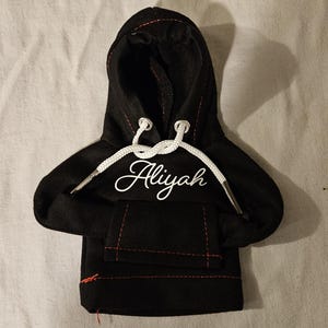 May include: Black miniature hoodie with a white drawstring and the name "Aliyah" in cursive script. The hoodie has a front pocket and red stitching details. The hood is up, and the drawstring is white.