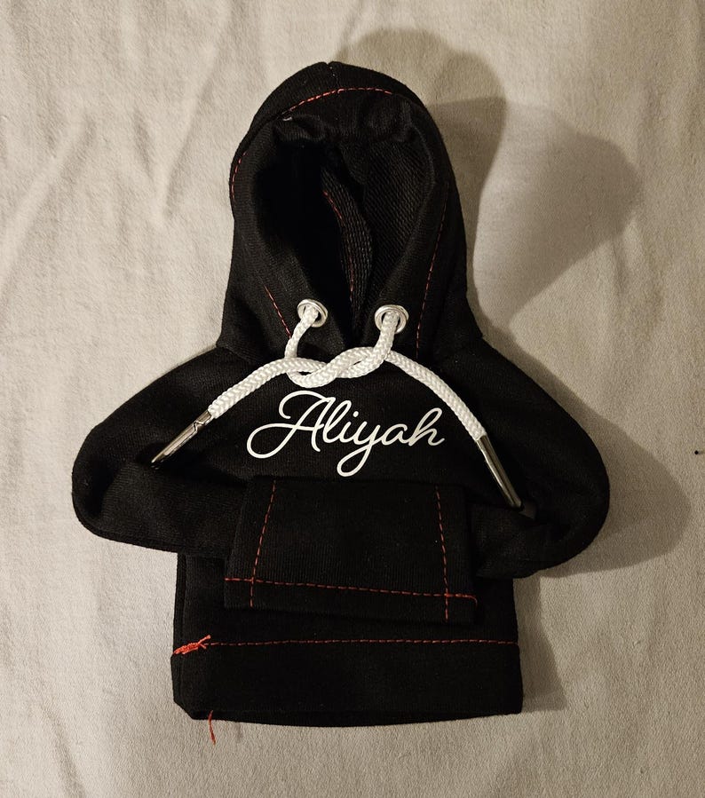 May include: Black miniature hoodie with a white drawstring and the name "Aliyah" in cursive script. The hoodie has a front pocket and red stitching details. The hood is up, and the drawstring is white.