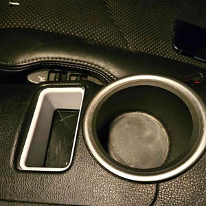 May include: Close-up of a black car interior with a cup holder and a small storage compartment. The cup holder is made of silver metal and has a black rubber insert. The storage compartment is made of black plastic and has a white trim.