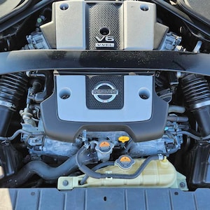 May include: Close-up view of a black Nissan V6 engine with a silver engine cover. The engine cover has the Nissan logo and the text "V6 VVVL" on it. The engine is in a car engine bay.