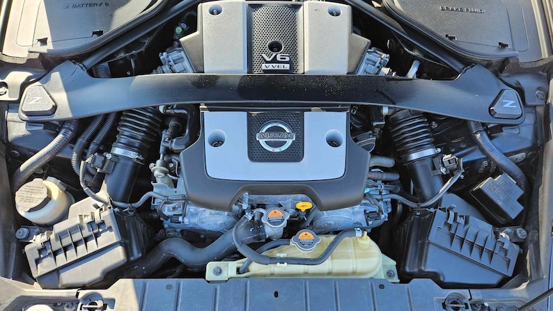May include: Close-up view of a black Nissan V6 engine with a silver engine cover. The engine cover has the Nissan logo and the text "V6 VVVL" on it. The engine is in a car engine bay.