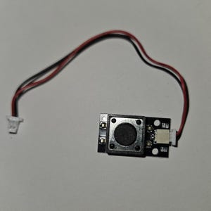 May include: A small black electronic component with a circular button in the center, surrounded by a square frame. It has a red and black wire attached, along with a white connector. The component is on a light gray surface.