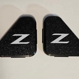May include: Two black triangular car emblems with a white "Z" on each. The emblems have a textured surface.