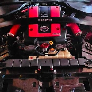 May include: A red and black engine compartment of a car with a red Nissan logo and the text "V6" on the engine cover.