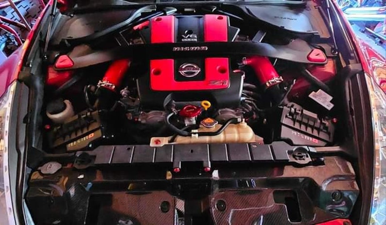 May include: A red and black engine compartment of a car with a red Nissan logo and the text "V6" on the engine cover.