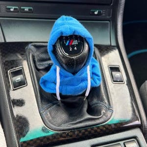 May include: A black gear shift knob with the BMW logo, covered by a miniature blue hoodie with white drawstrings. The shift knob is surrounded by a black leather console with various buttons and switches.