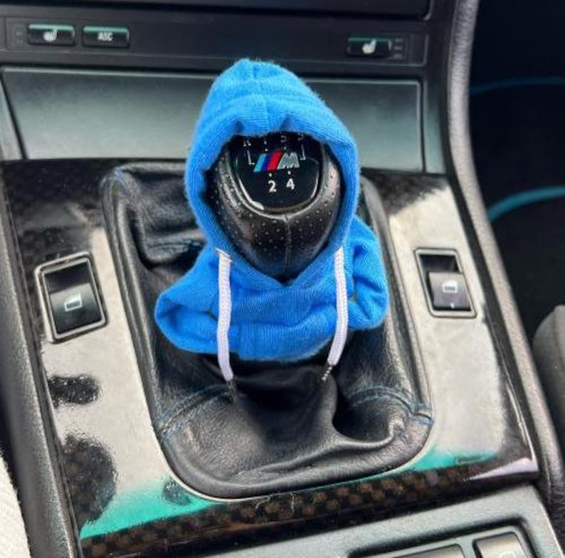 May include: A black gear shift knob with the BMW logo, covered by a miniature blue hoodie with white drawstrings. The shift knob is surrounded by a black leather console with various buttons and switches.