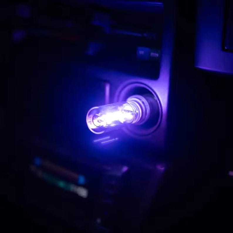 May include: A glowing, cylindrical USB car charger with a bright blue light. The charger is plugged into a car's dashboard, illuminating the surrounding area with a cool, purple hue. The image is dark, focusing on the charger and its emitted light.