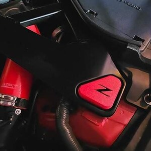 May include: Close-up of a red engine compartment with a black plastic cover and a red triangular emblem with a white "Z" inside. The emblem is attached to the cover.
