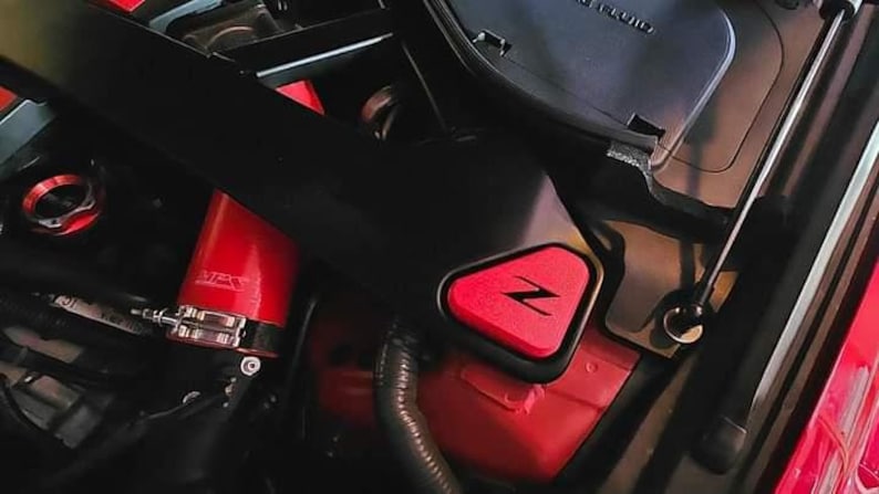 May include: Close-up of a red engine compartment with a black plastic cover and a red triangular emblem with a white "Z" inside. The emblem is attached to the cover.
