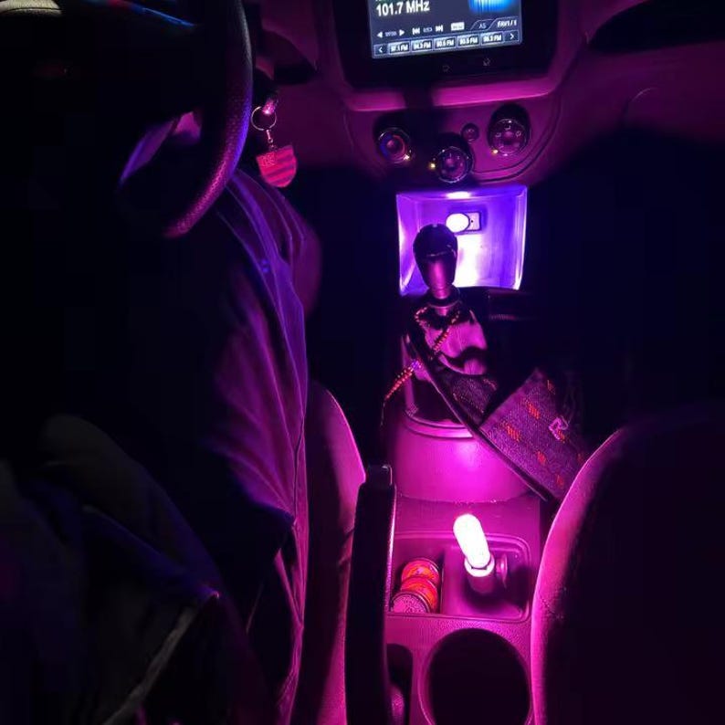 May include: Interior car shot illuminated with purple lighting. The gear shift, center console, and dashboard are visible. A small light is in the cup holder. The radio displays 101.7 MHz. A steering wheel is in the upper left corner.