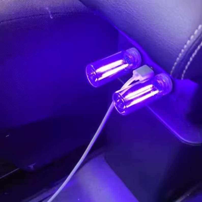 May include: Two small, clear glass vials with screw-on caps are illuminated with a bright purple light. A white cable connects the vials, suggesting a charging or lighting function. The vials are positioned in a car's interior.