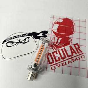 May include: A clear USB drive with orange internal components is displayed next to two stickers. One sticker features a cartoon ninja turtle design with Japanese text. The other sticker has a red graphic of a race car driver and the words "OCULAR DYNAMIC".