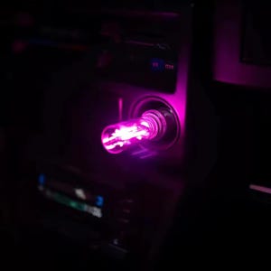 May include: A glowing pink light bulb inserted into a car's cigarette lighter port. The bulb emits a bright, vibrant pink light, illuminating the surrounding area. The car's dashboard and interior are visible in the background.