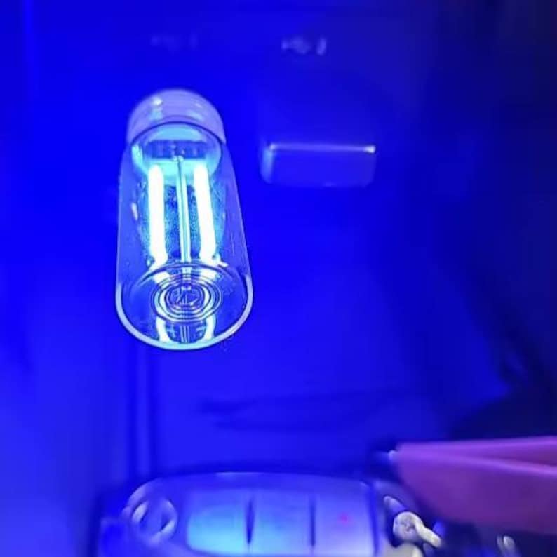 May include: A clear glass tube with a light blue glow, possibly a UV lamp, against a dark blue background. The tube has a rounded bottom and a silver cap. The image is brightly lit with a focus on the tube.